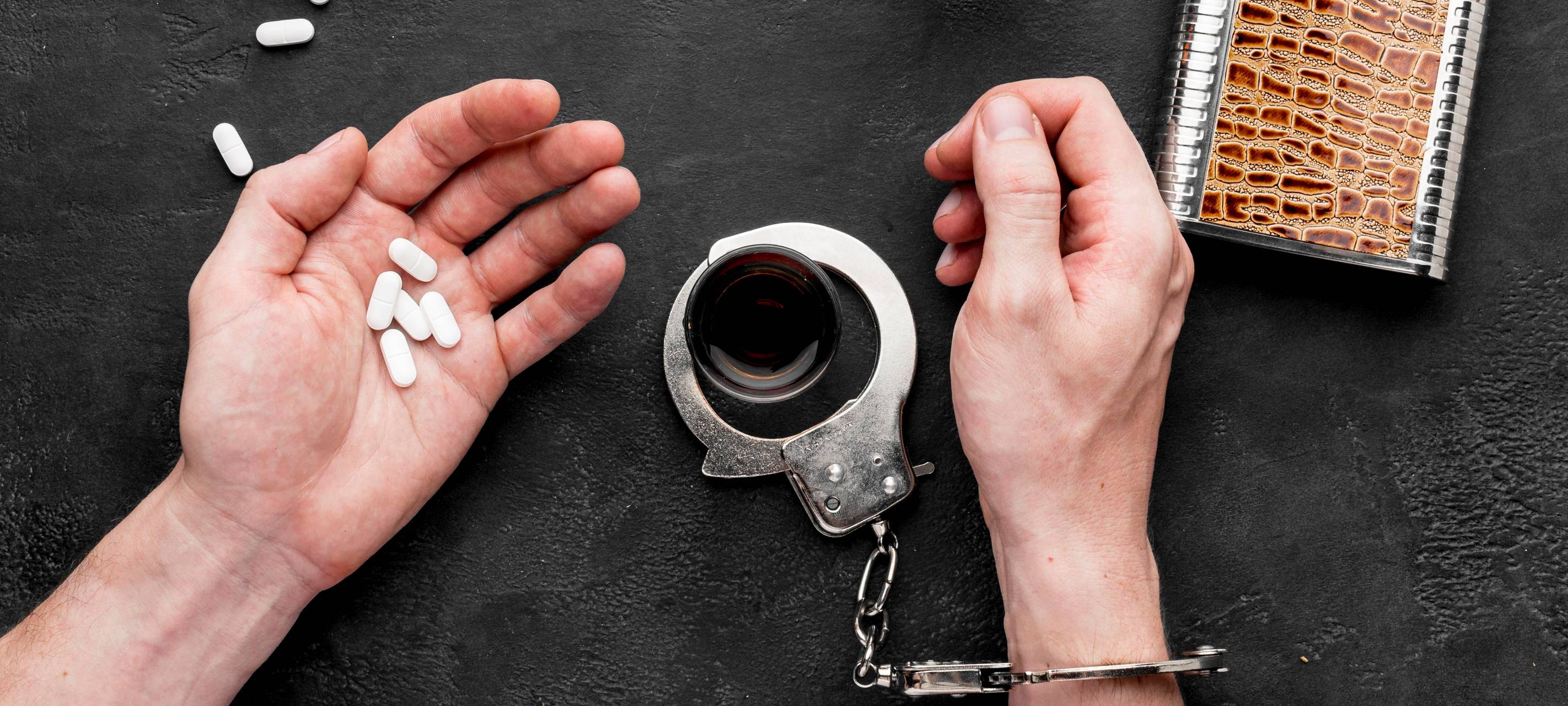 Männerhände mit Pillen in der Hand, Drogen und Handschellen auf einem Tisch